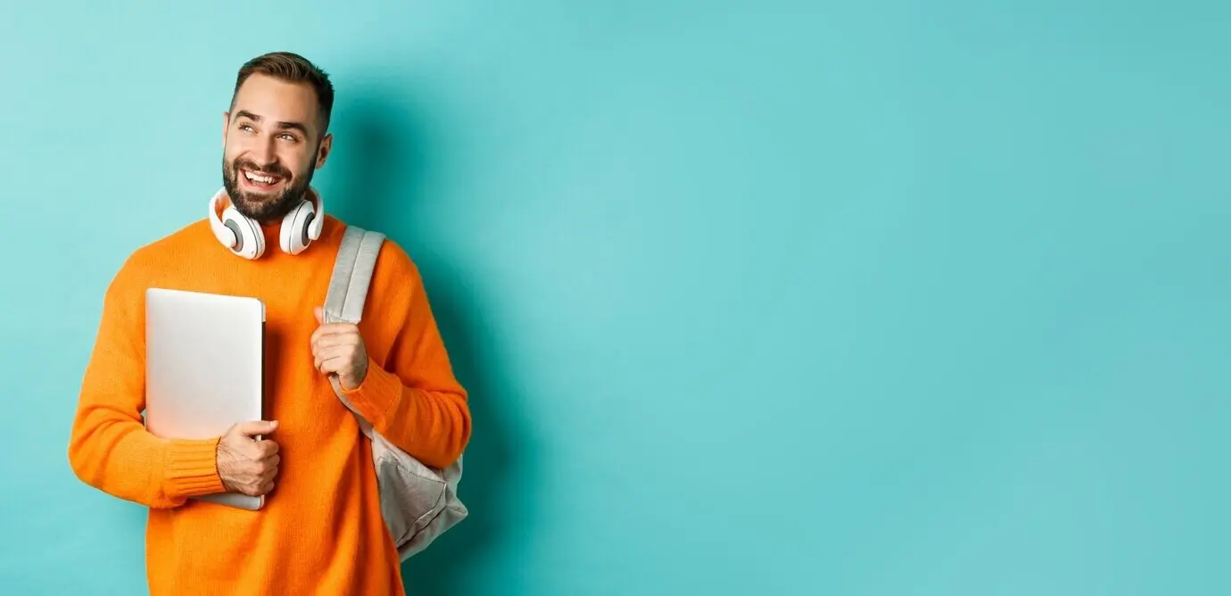 Ein fröhlicher Mann mit Rucksack und Kopfhörern hält einen Laptop, lächelt, blickt nachdenklich nach links und steht.